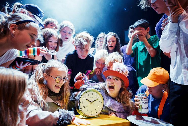 A group of children performing Alice in Wonderland. They are staring at an old fashioned alarm clock in amazement.